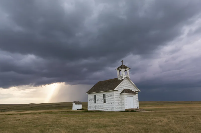 Small white clapboard church standing alone in a grassy prairie field under a stormy sky with a sunbeam breaking through the clouds