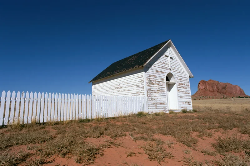 Small weathered white wooden chapel with picket fence in arid desert landscape under clear blue sky