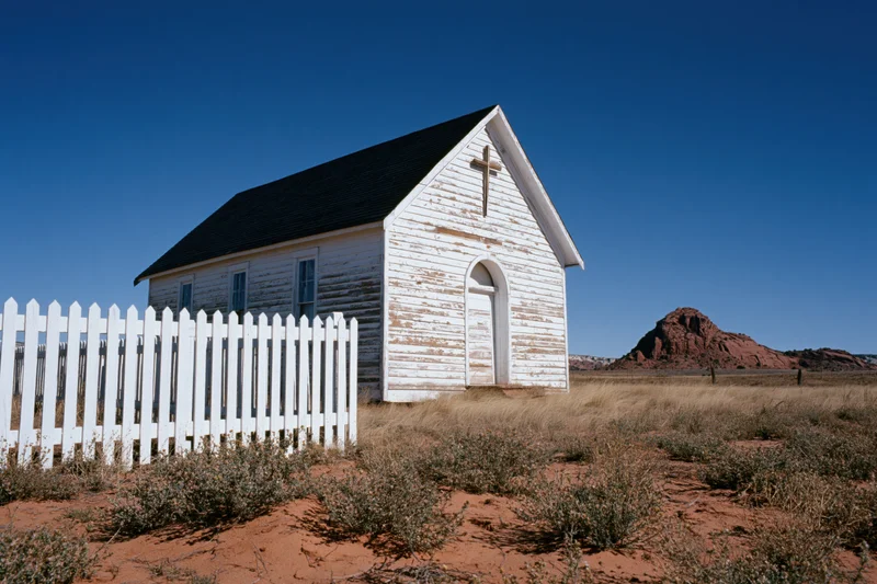 Weathered white wooden chapel with picket fence in desert landscape under clear blue sky