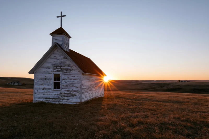 Small weathered white wooden church with cross on prairie at sunset