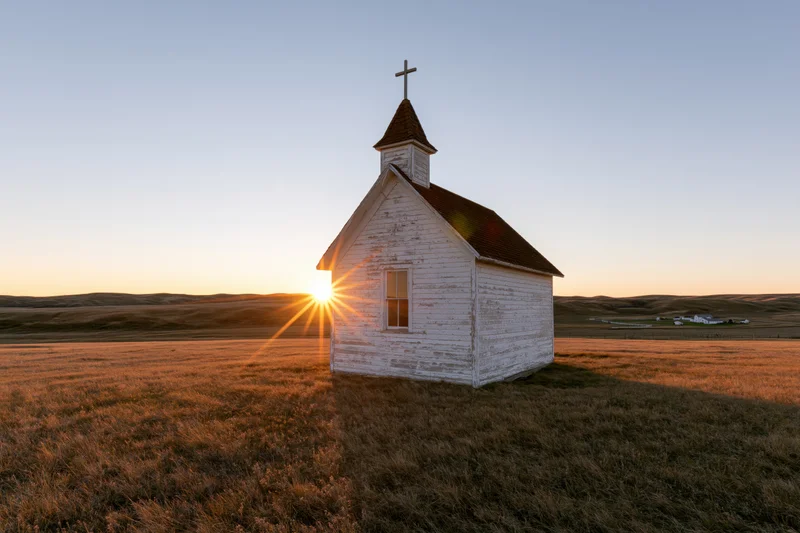 Small weathered white wooden church with cross on prairie at sunset