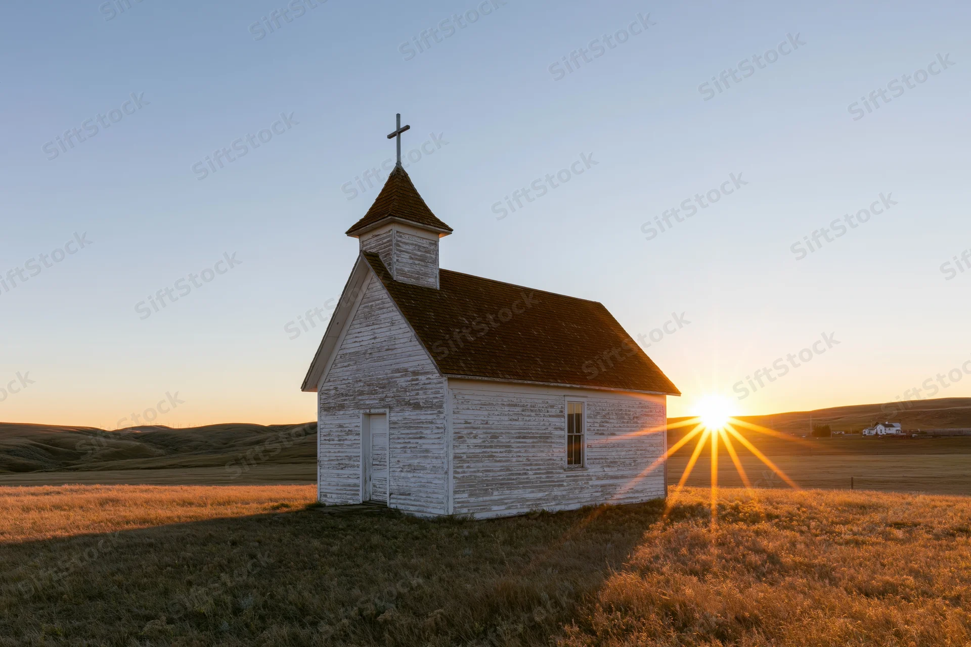 Small weathered white wooden church with cross on prairie at sunset