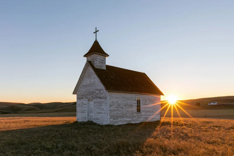 Small weathered white wooden church with cross on prairie at sunset
