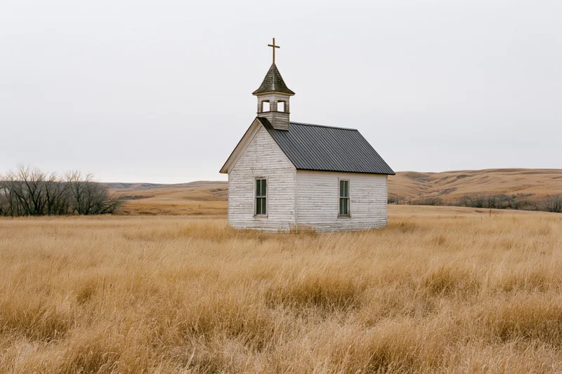 Small weathered wooden church with steeple and cross standing in a dry grassy prairie field