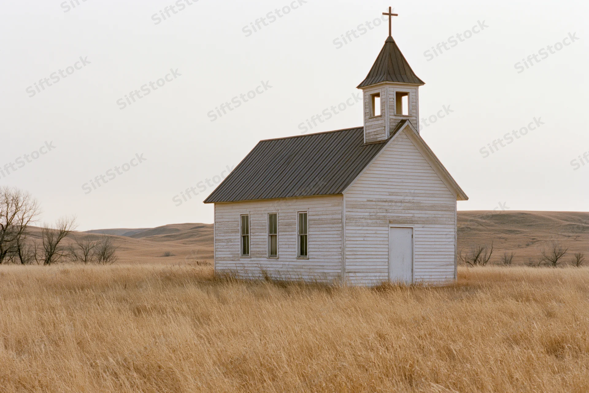 Small weathered wooden church with steeple and cross standing in a dry grassy prairie field