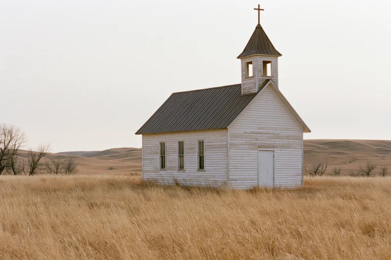 Small weathered wooden church with steeple and cross standing in a dry grassy prairie field