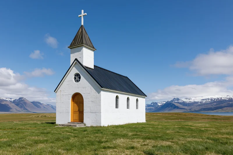 Small white church with wooden arched door standing in a grassy field with mountains and blue sky in the background