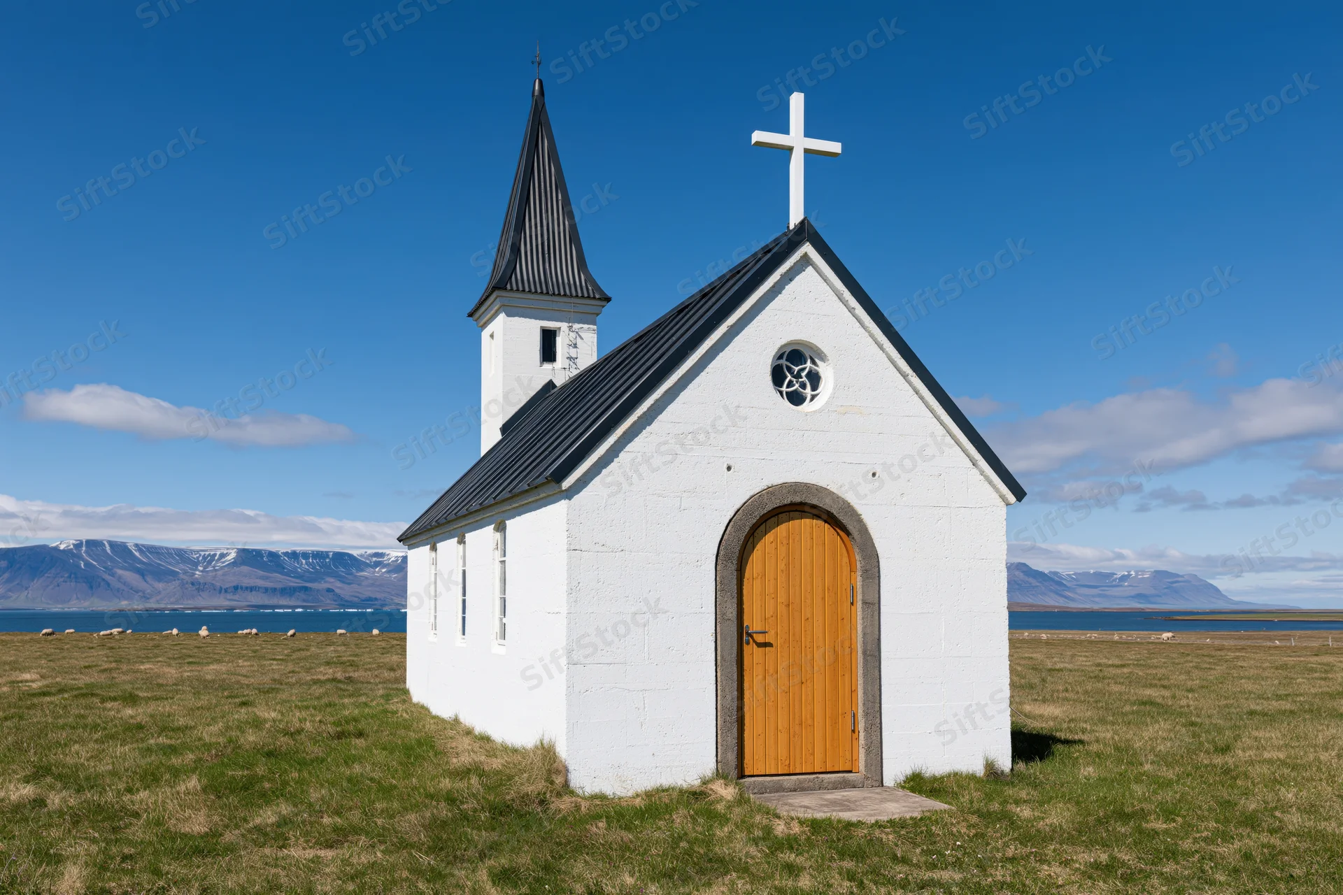 Small white icelandic church with arched wooden door and cross on grassy field near mountains and fjord