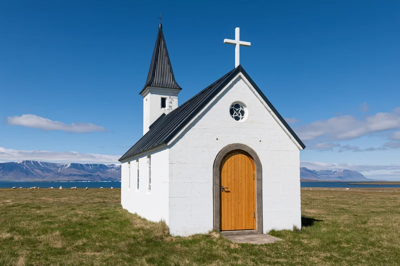 Small white icelandic church with arched wooden door and cross on grassy field near mountains and fjord