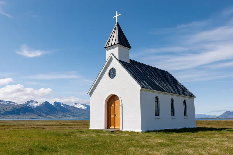 Small white icelandic church with wooden arched door in grassy field near snow-capped mountains