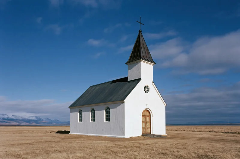 Small white church with dark metal steeple in open rural field under blue sky