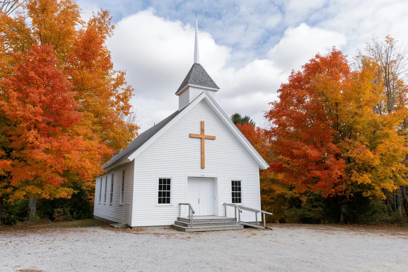 Small white baptist church with wooden cross on facade surrounded by autumn trees in rural setting