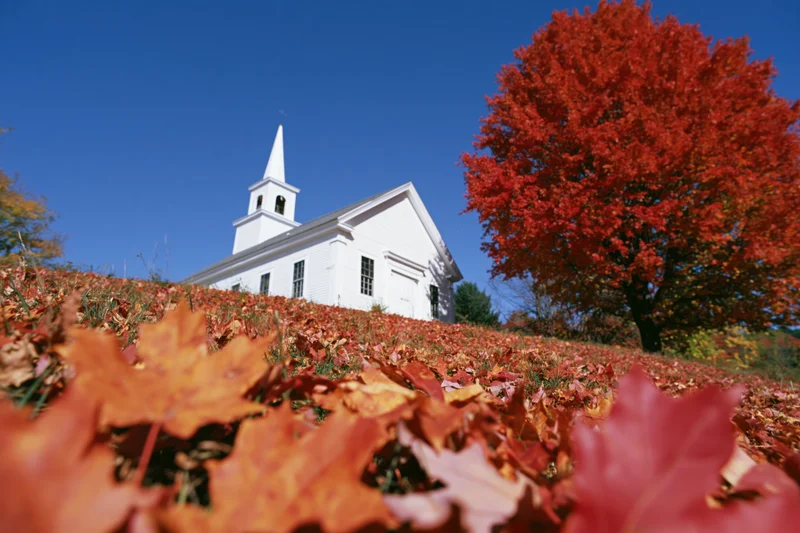 White clapboard church with steeple on a hill surrounded by autumn foliage under a clear blue sky