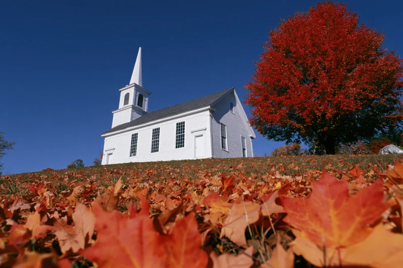 White clapboard church with steeple on a hill surrounded by autumn leaves under a clear blue sky