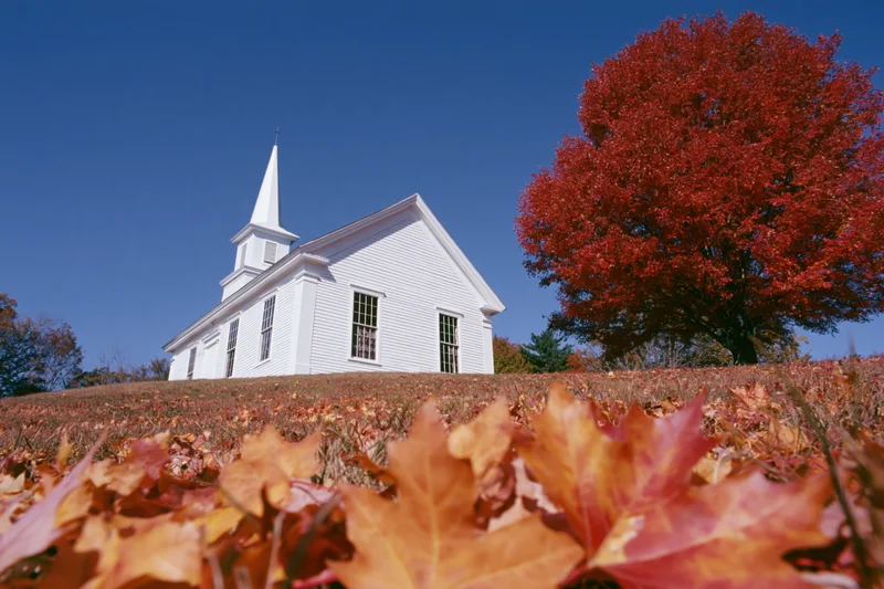 White clapboard church with steeple on a hill surrounded by autumn foliage under a clear blue sky