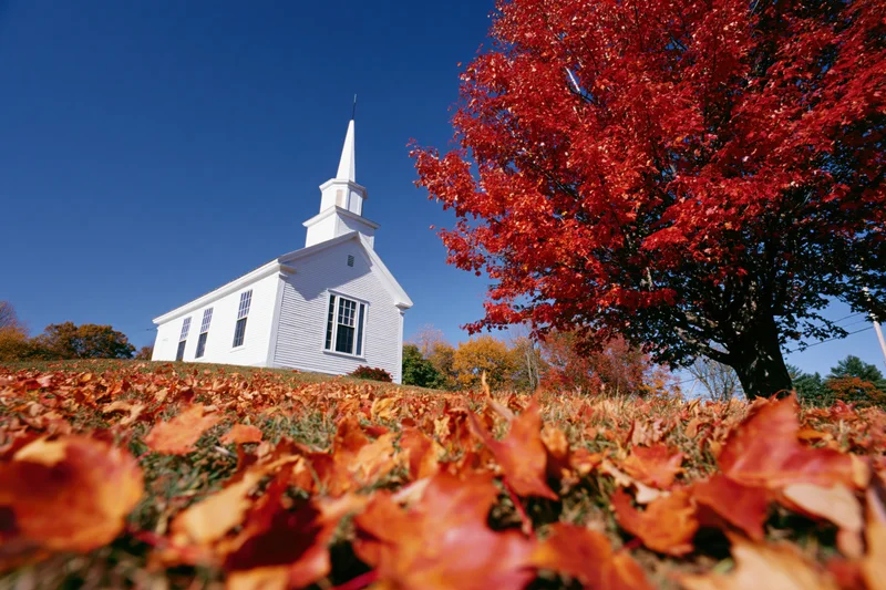 Small white clapboard church with steeple surrounded by red autumn foliage under a clear blue sky