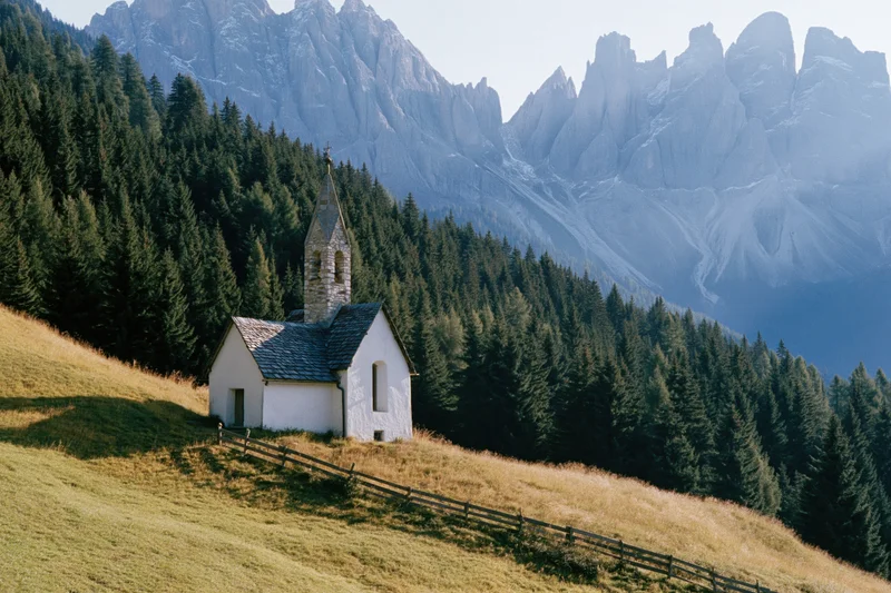 Small white alpine chapel with stone bell tower on grassy hillside surrounded by pine forest and mountains