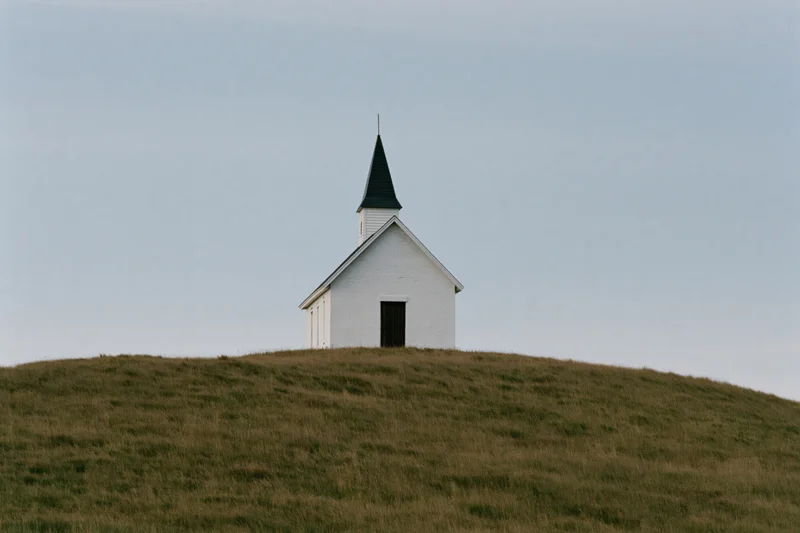 Small white chapel with dark steeple on grassy hill under overcast sky