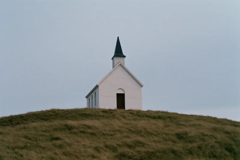 Small white chapel with pointed steeple on a grassy hill under overcast sky