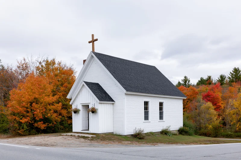 Small white clapboard church with wooden cross on roof surrounded by autumn foliage along a rural roadside