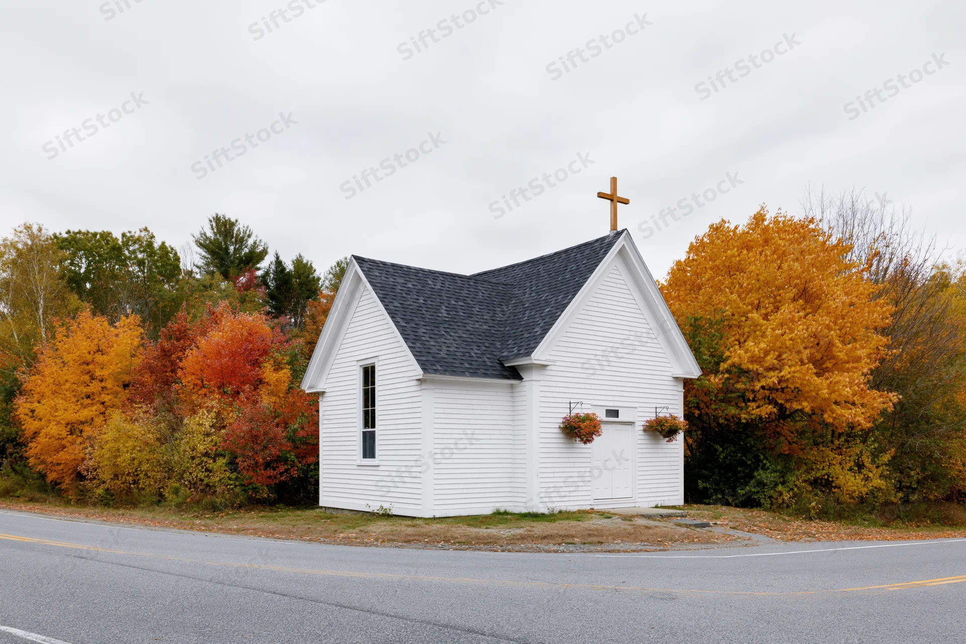 Small white clapboard church with wooden cross on roof beside autumn trees along rural road