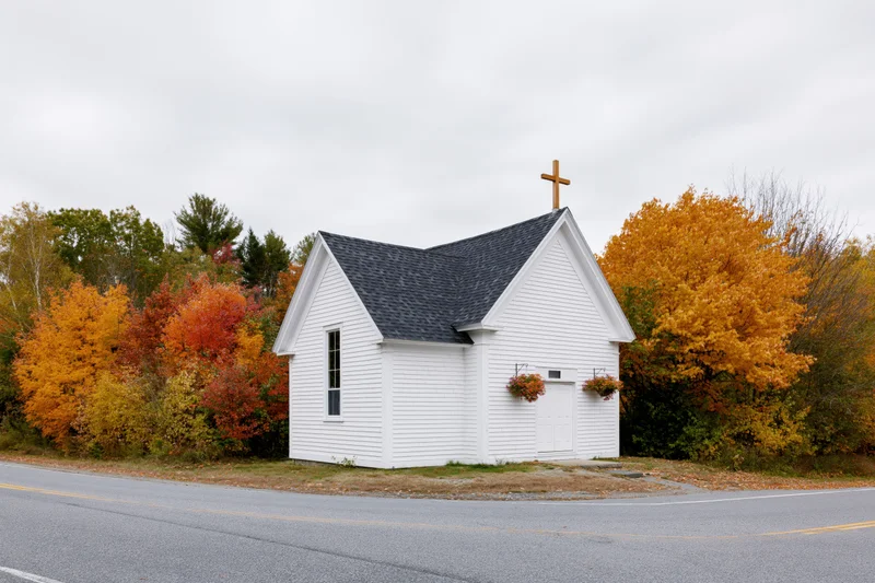 Small white clapboard church with wooden cross on roof beside autumn trees along rural road