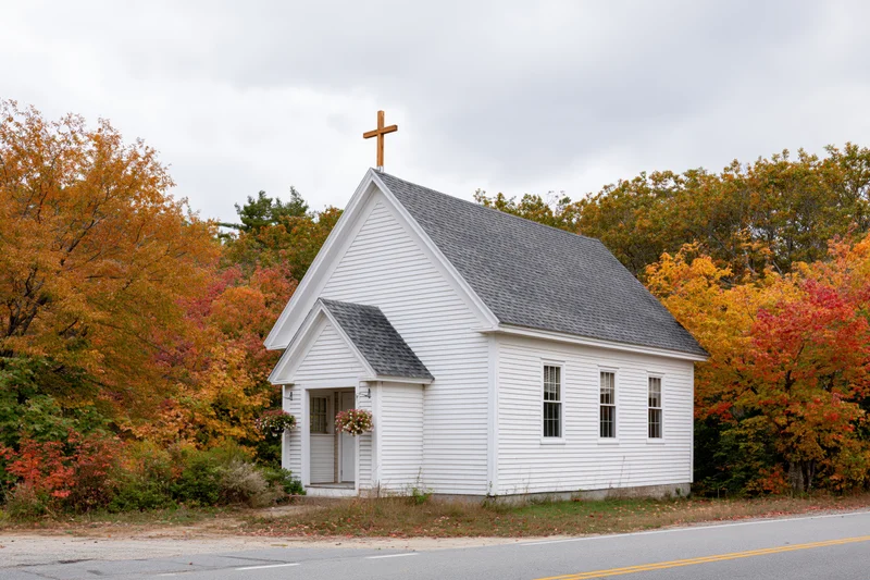 Small white clapboard church with wooden cross on roof beside autumn trees along rural road