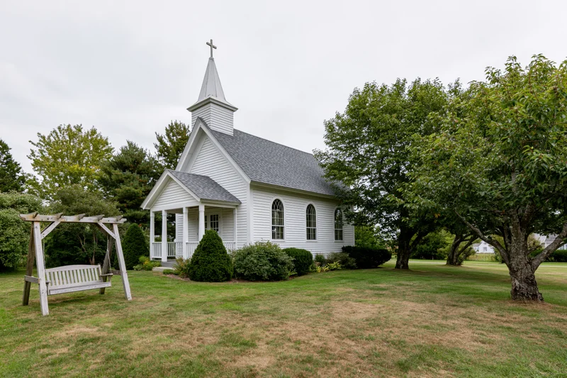 Small white clapboard chapel with cross-topped steeple and porch surrounded by green lawn and trees