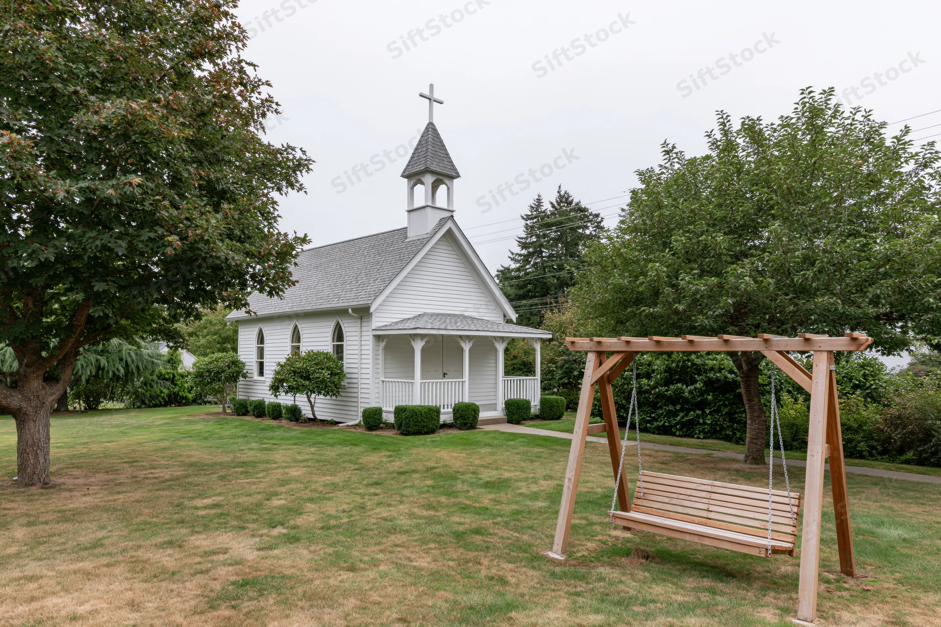 Small white clapboard chapel with cross-topped steeple and porch, surrounded by grassy lawn and trees, with wooden swing in foreground