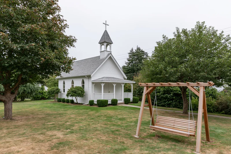Small white clapboard chapel with cross-topped steeple and porch, surrounded by grassy lawn and trees, with wooden swing in foreground
