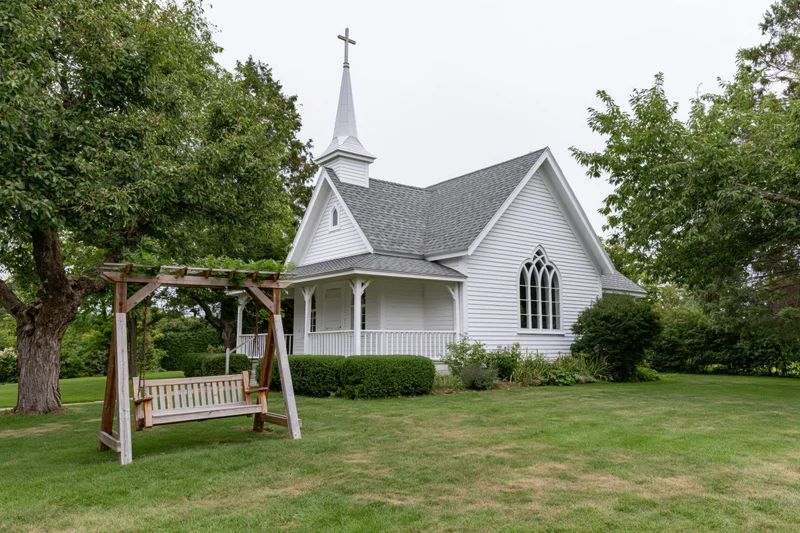 Small white clapboard chapel with cross-topped steeple and porch surrounded by green lawn and trees