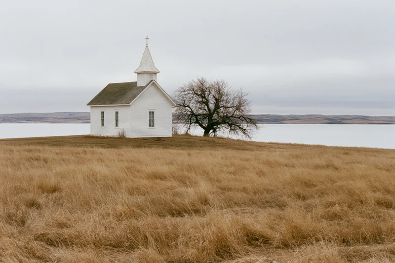 Small white clapboard church with steeple and cross on a grassy hill overlooking water