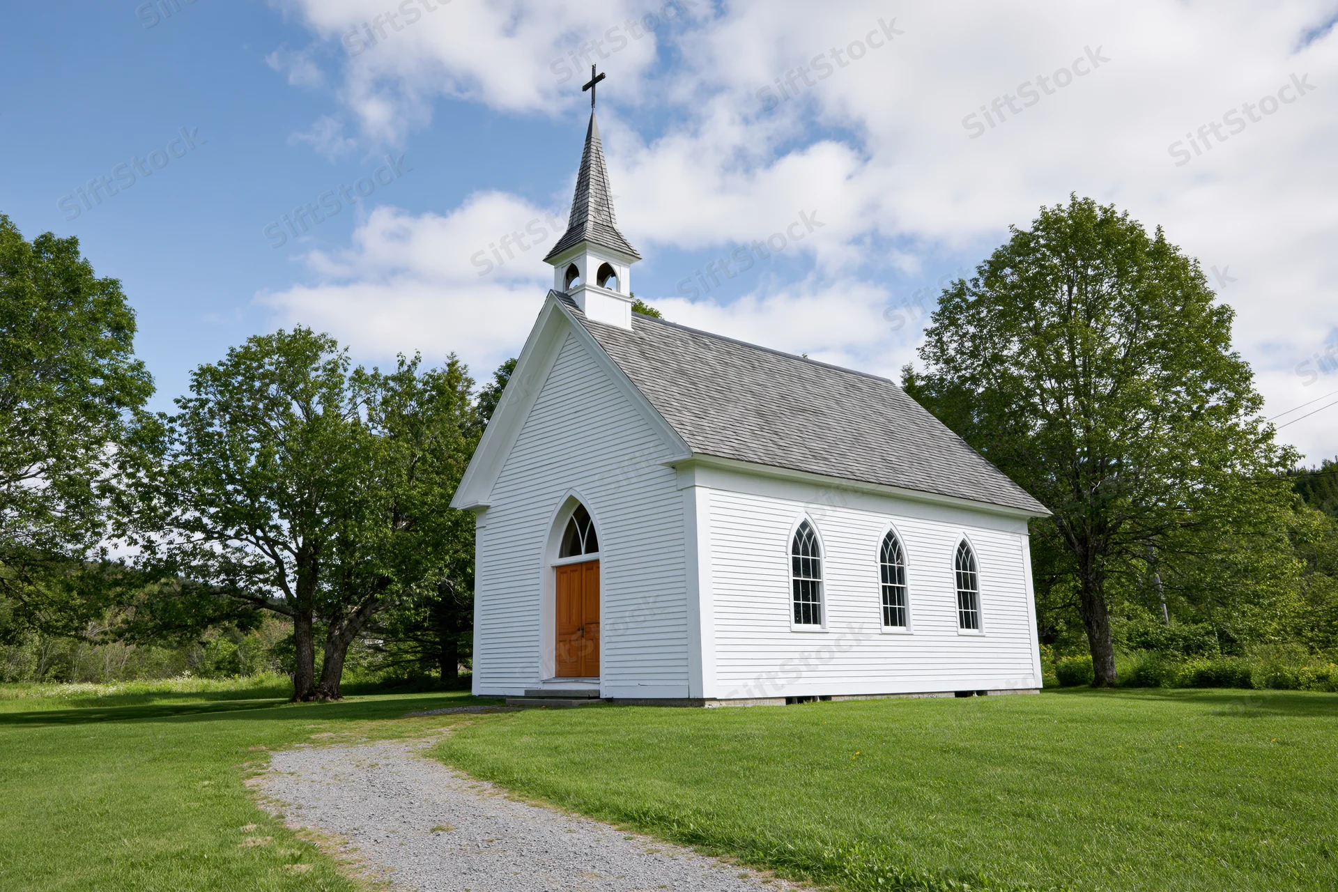 Small white clapboard church with pointed steeple and brown door on grassy field surrounded by trees under blue sky