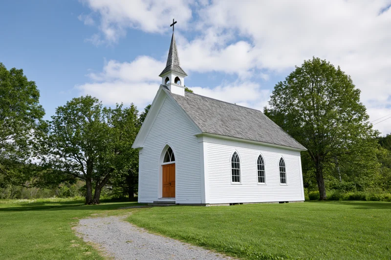 Small white clapboard church with pointed steeple and brown door on grassy field surrounded by trees under blue sky