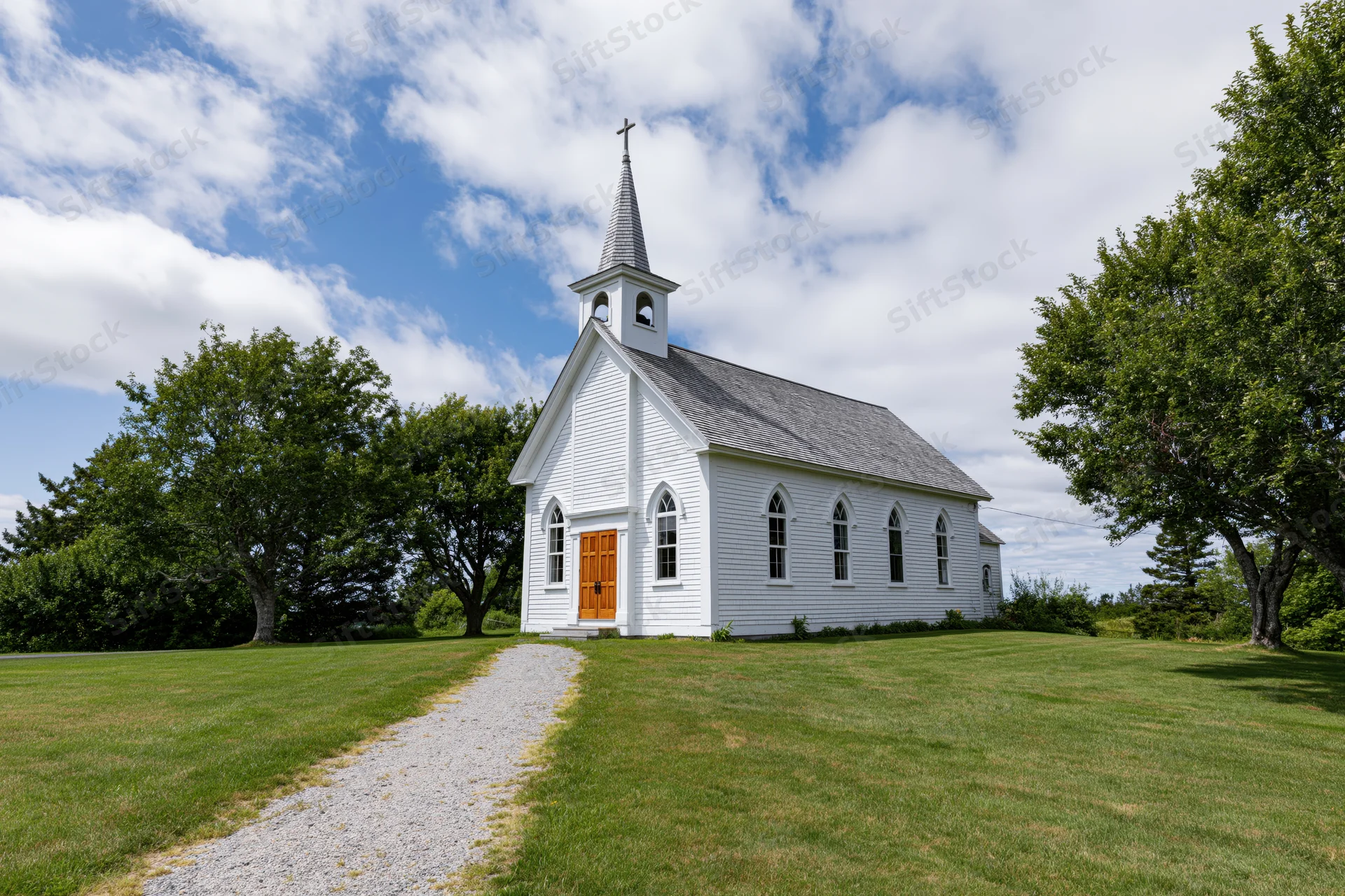 Small white clapboard church with pointed steeple and brown door on grassy lawn surrounded by trees under blue sky with clouds