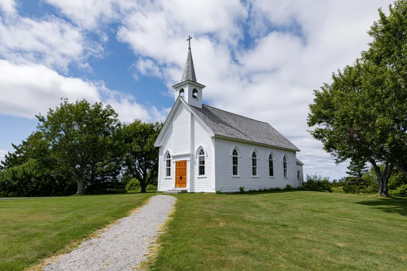 Small white clapboard church with pointed steeple and brown door on grassy lawn surrounded by trees under blue sky with clouds