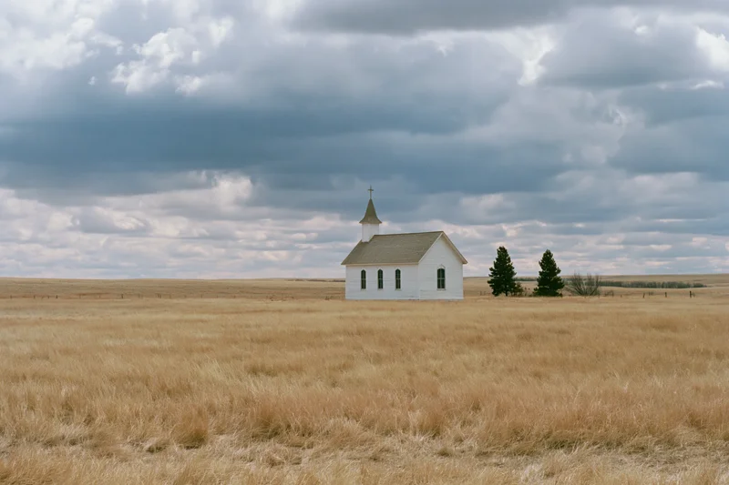 Small white clapboard church with steeple and cross standing alone in a dry grass field under cloudy sky