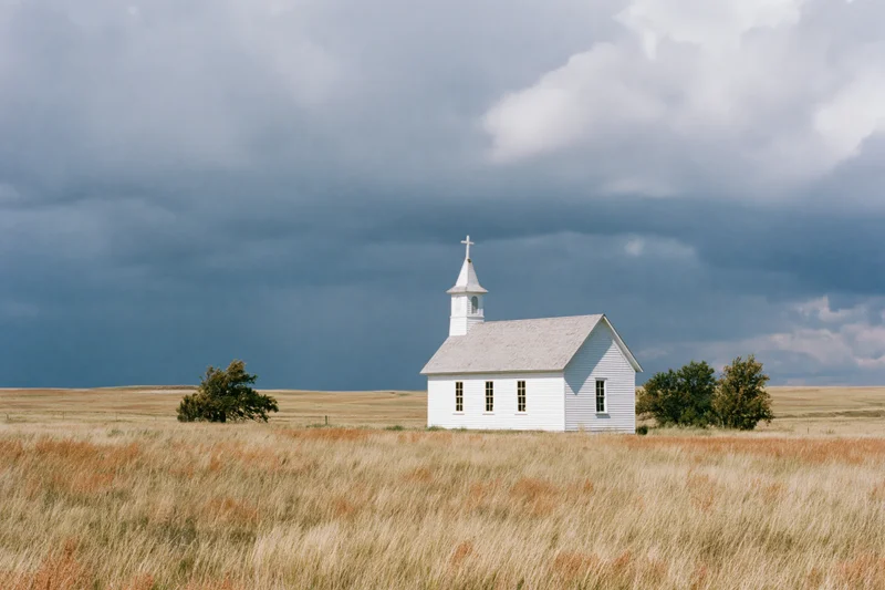 Small white clapboard church with steeple and cross standing in a grassy field under stormy sky