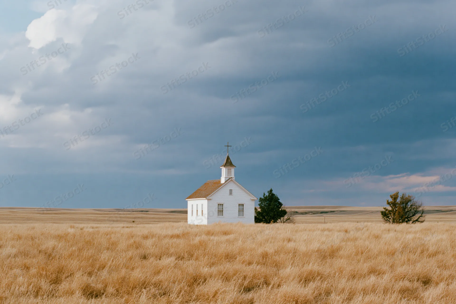 Small white clapboard church with steeple and cross standing alone in a golden grass field under cloudy sky