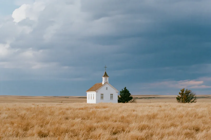 Small white clapboard church with steeple and cross standing alone in a golden grass field under cloudy sky