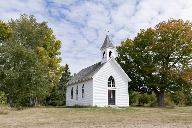 Small white clapboard church with steeple and cross in rural field surrounded by autumn trees