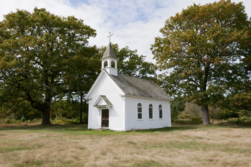 Small white clapboard church with steeple and cross in a rural field surrounded by trees