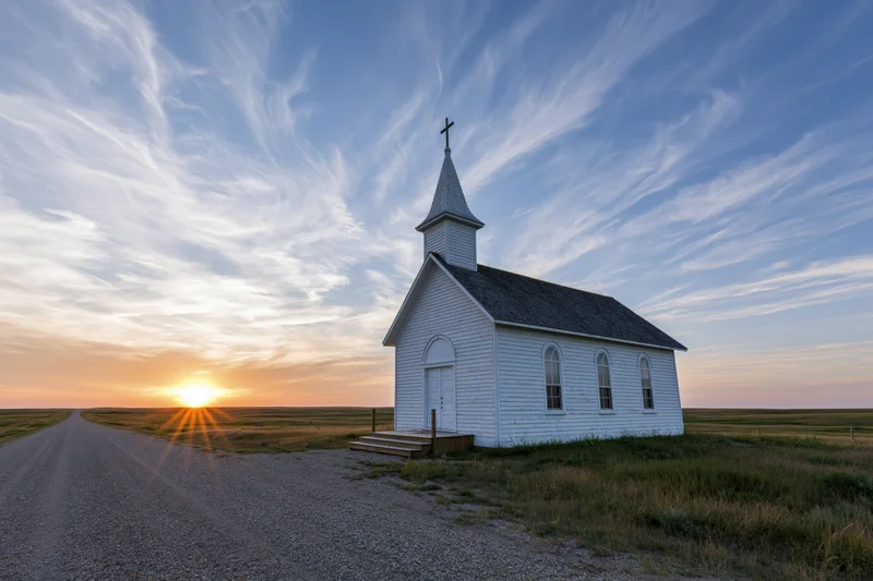 Small white clapboard church with steeple and cross at sunset on a gravel road in open prairie field