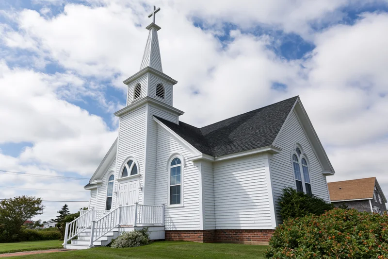 Small white clapboard church with tall steeple and cross under blue sky with clouds