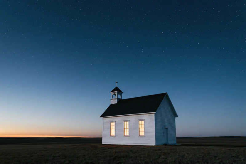 Small white clapboard church with bell tower and cross under starry night sky on prairie field at twilight
