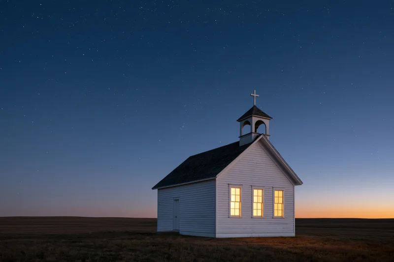 Small white clapboard church with bell tower and cross illuminated at twilight on prairie field