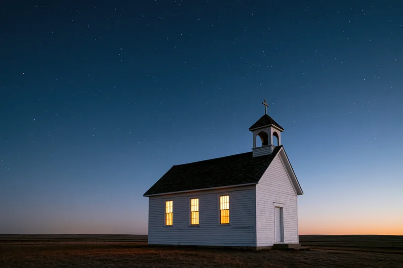 Small white clapboard church with bell tower and cross under starry night sky on prairie field at twilight