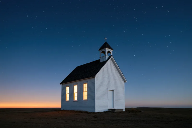 Small white clapboard church with bell tower and cross under starry sky on prairie at twilight