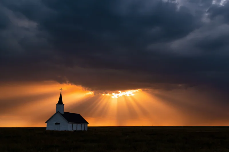 Small white country church silhouetted against dramatic sunset sky with sun rays breaking through clouds over prairie field