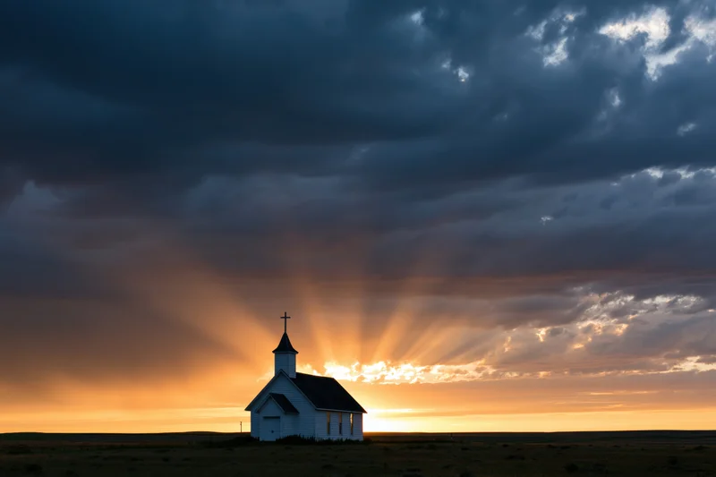 Small white country church silhouetted against dramatic sunset sky with sun rays breaking through clouds